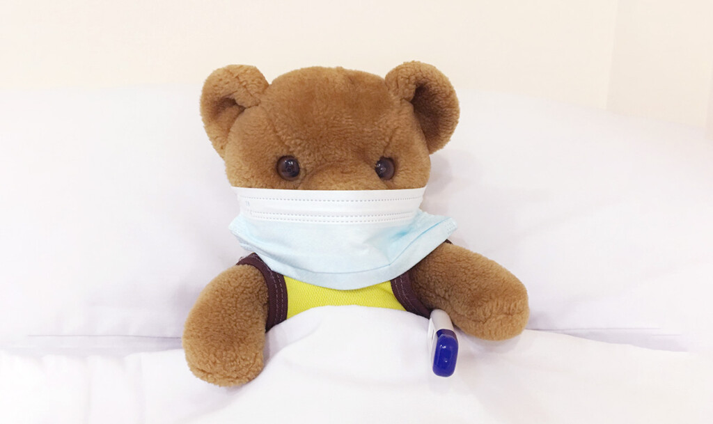 A teddy bear wearing a medical face mask tucked in bed